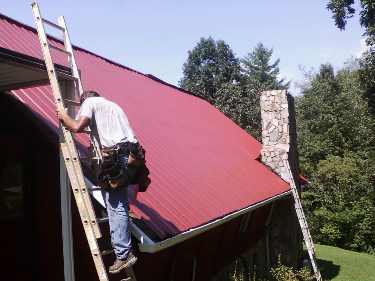 Roofer on a ladder beside a red metal roof and stone chimney — Soffit & Fascia Repair by Roofing Brothers in Mckeesport